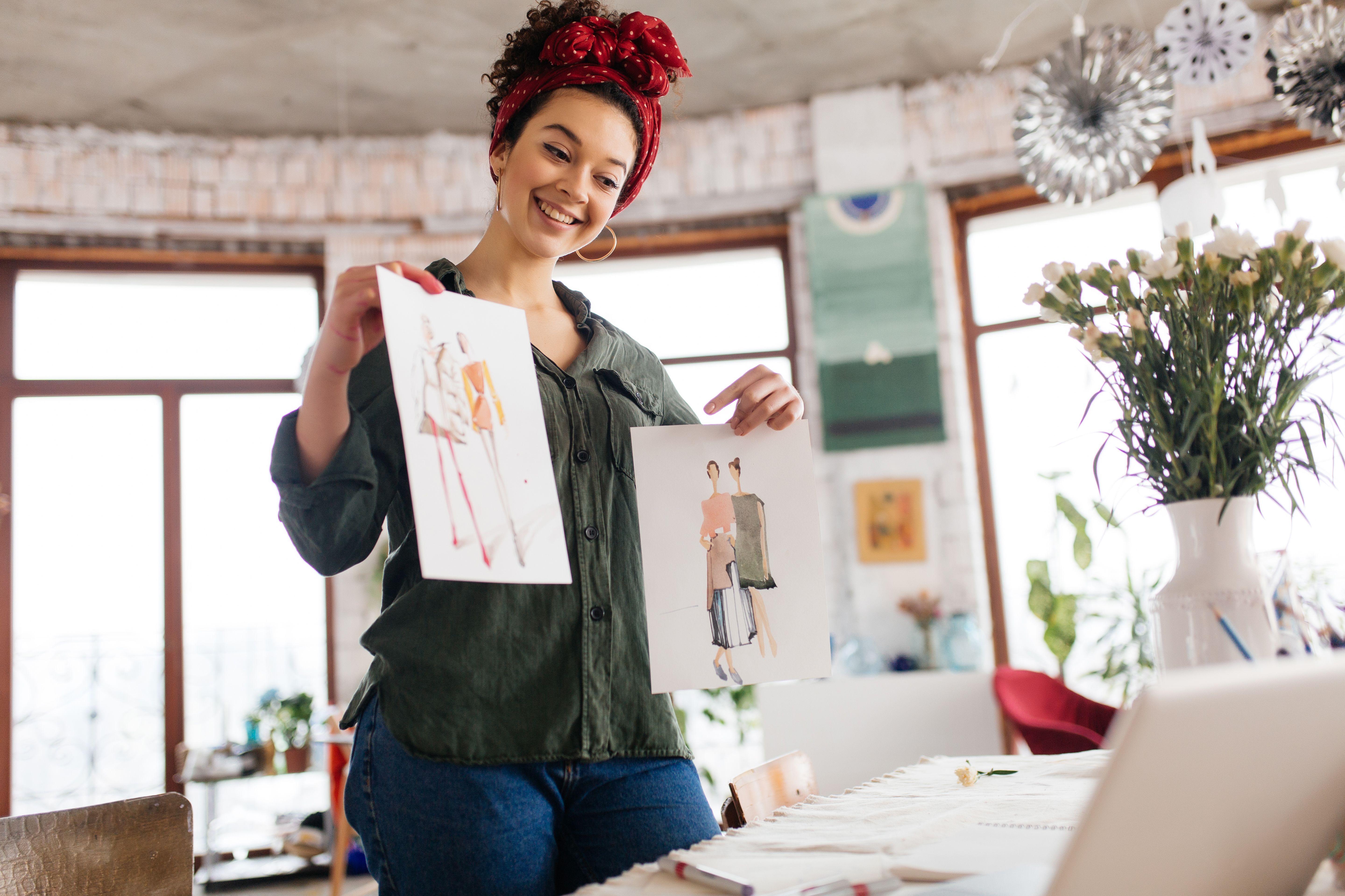 young-joyful-woman-with-dark-curly-hair-standing-near-table-happily-showing-fashion-sketches-laptop-while-spending-time-modern-cozy-workshop-with-big-windows.jpg